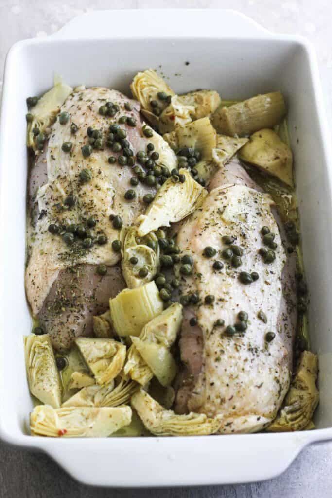 raw chicken breasts with capers and artichokes in a white baking dish
