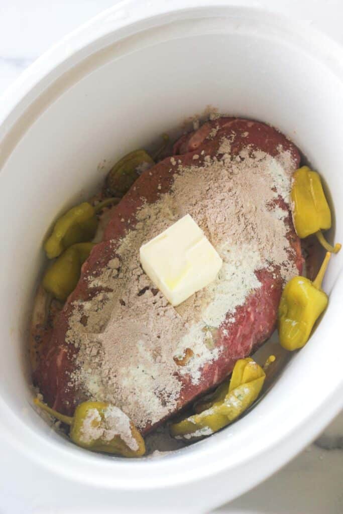 peperoncinis and seasoning on top a large piece of beef in crock pot