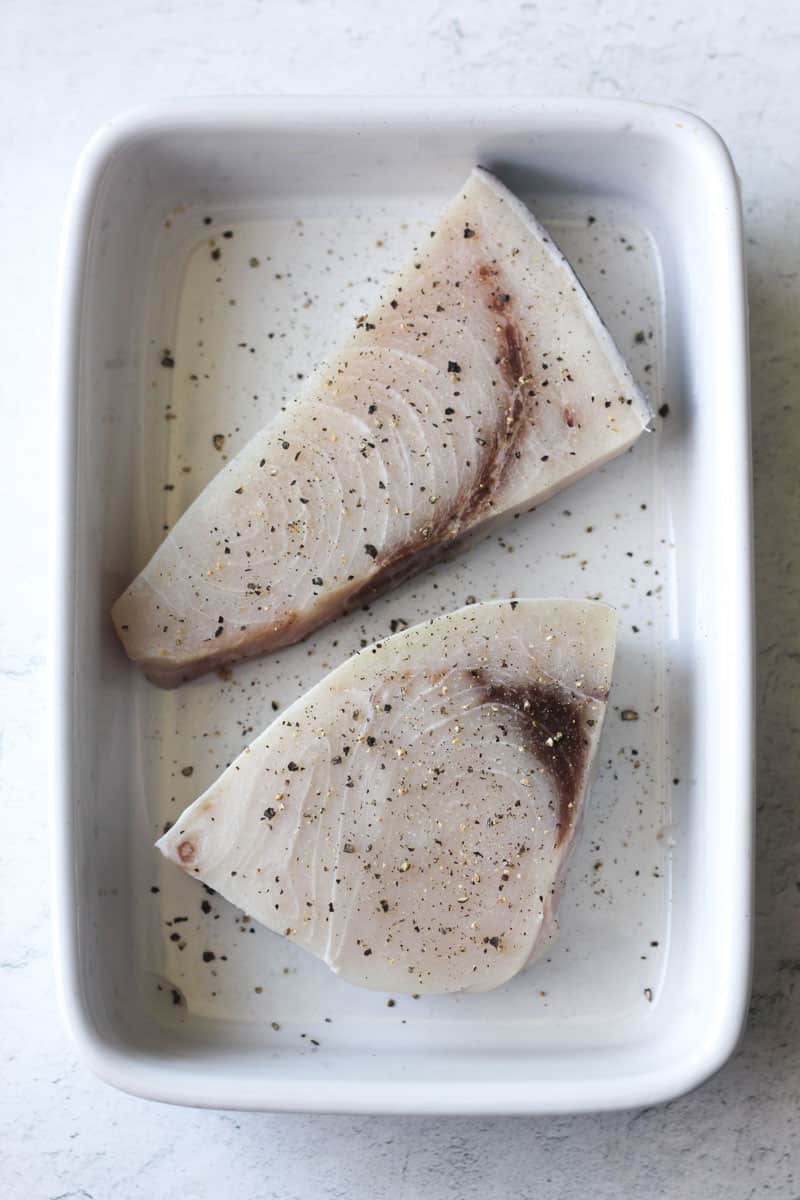 two raw swordfish steaks covered with black pepper in the white baking dish