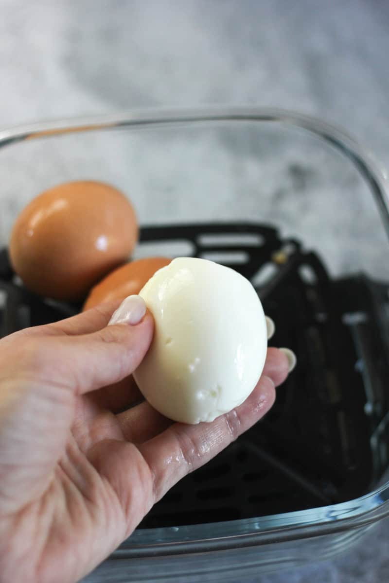 Close-up of a hand holding a peeled, cooked egg over a glass container with raw eggs in the backgrou.