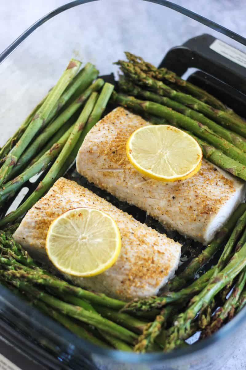 Baked fish fillets with lemon slices served alongside roasted asparagus in a glass dish.