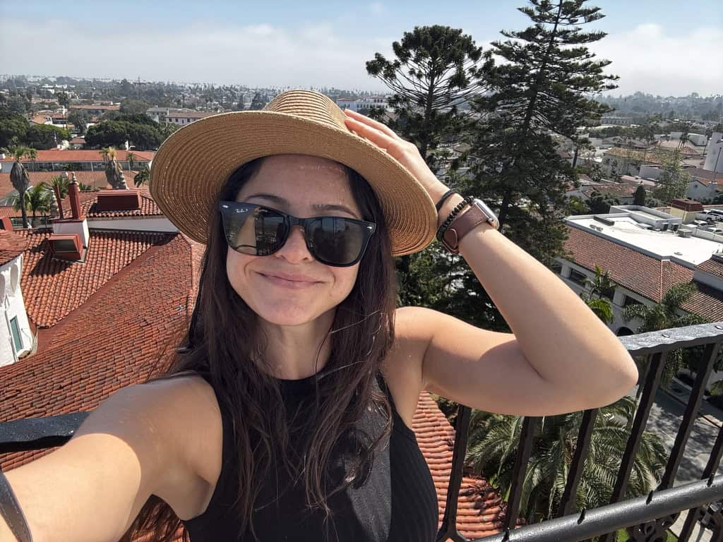 Woman wearing a hat and sunglasses on balcony overlooking cityscape.