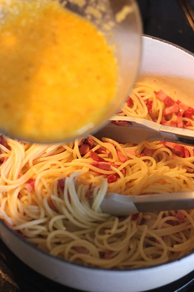 Pouring beaten eggs into a pan of cooked spaghetti with bacon bits.