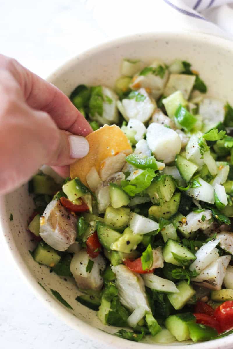 Hand adding a cracker to a bowl of chicken and cucumber salad with herbs.