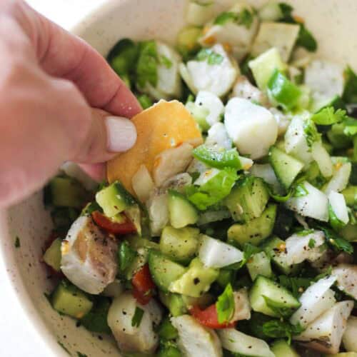 Hand adding a cracker to a bowl of chicken salad with cucumbers and tomatoes.
