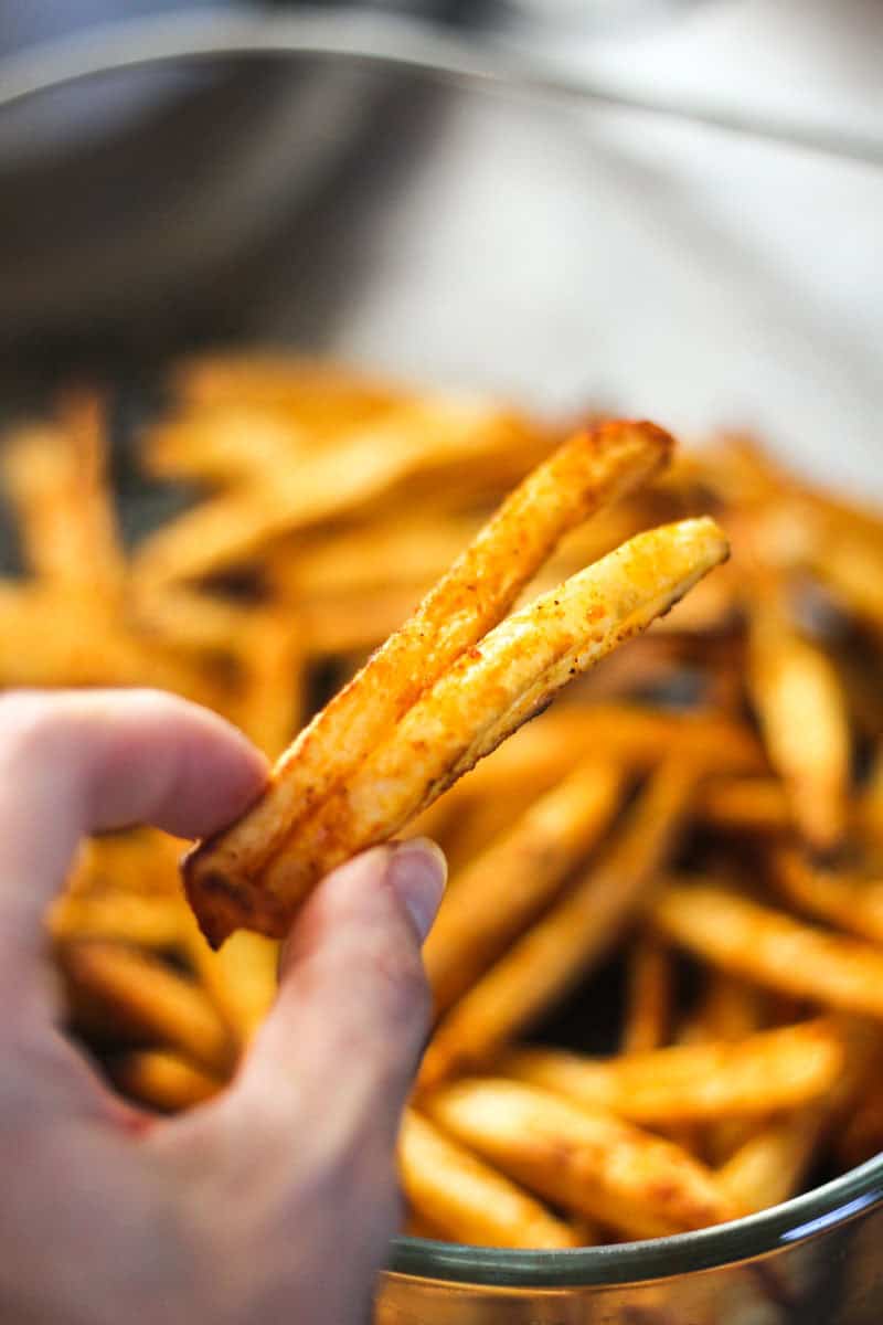 Golden crispy french fries held between fingers, bowl of fries in background.